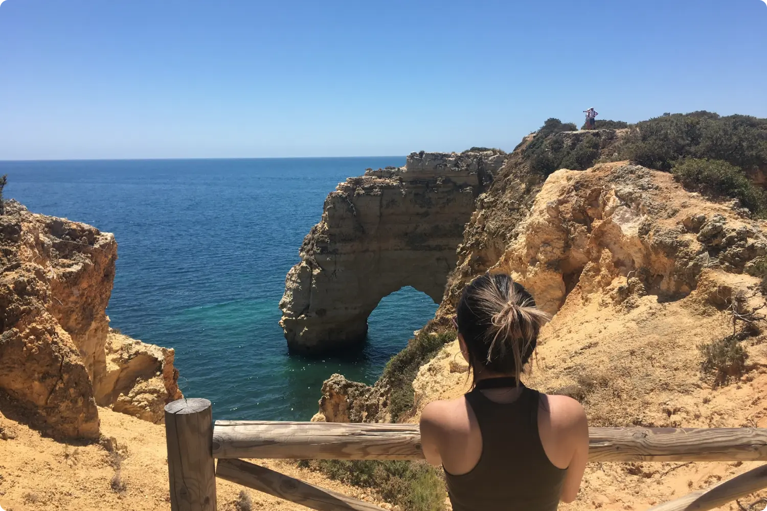 A woman stands at a wooden railing, overlooking a rocky cliff and ocean arch under a clear blue sky