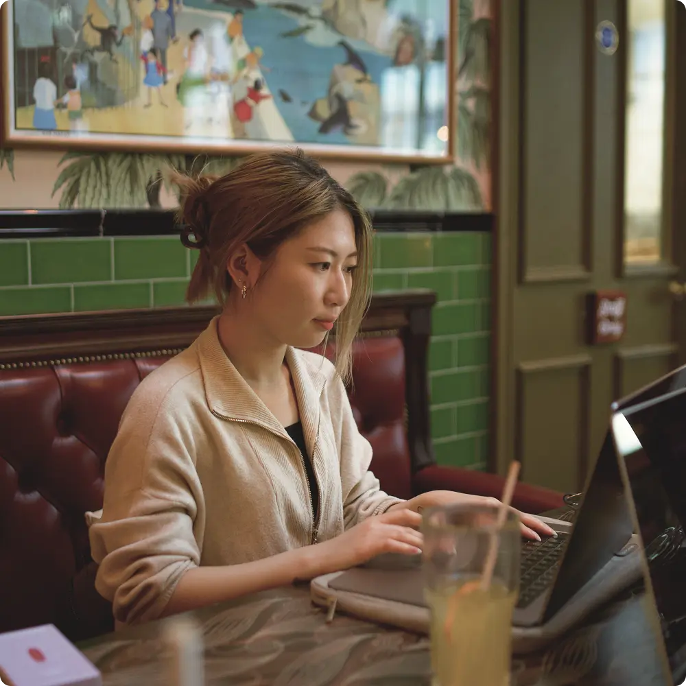 A woman works on a laptop at a cafe with a relaxed, focused expression. The setting features green tiles, a vibrant wall painting, and a glass of juice.
