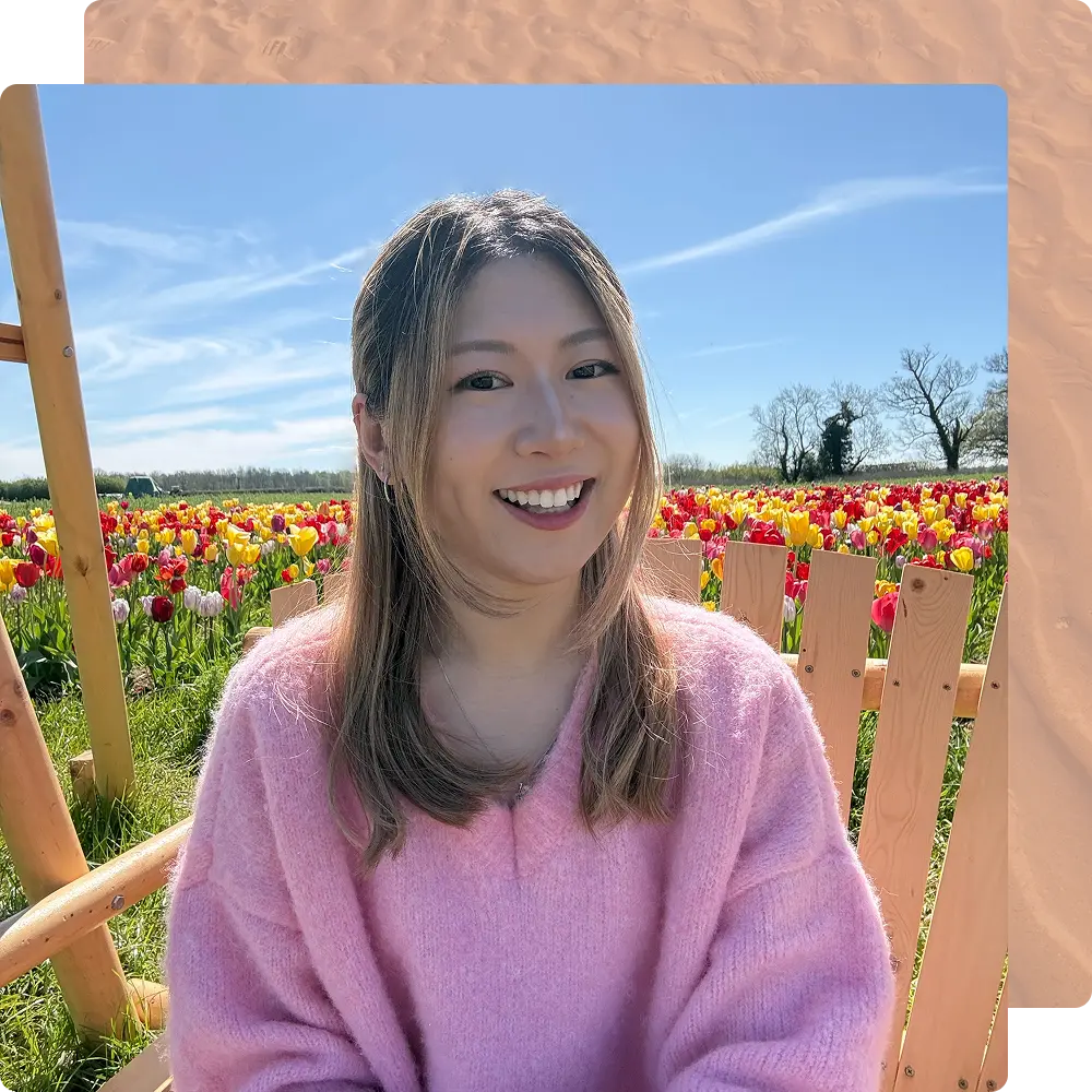 A person in a pink sweater smiles while sitting on a wooden bench amid a vibrant tulip field under a clear blue sky.
