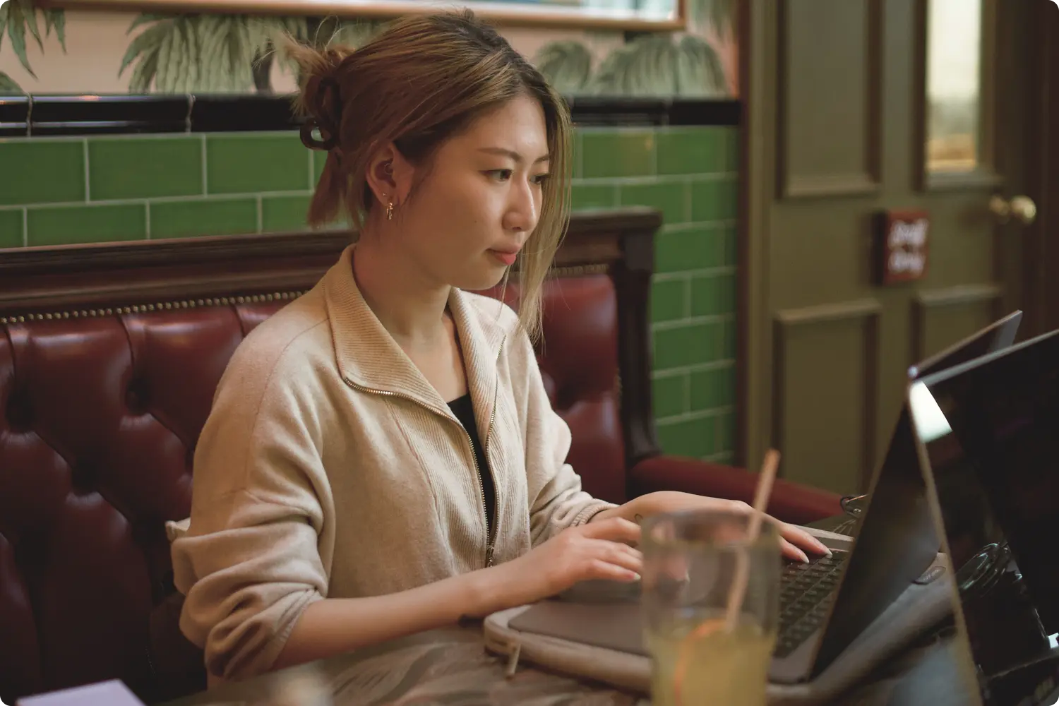 A woman works on a laptop at a cafe with a relaxed, focused expression. The setting features green tiles, a vibrant wall painting, and a glass of juice.
