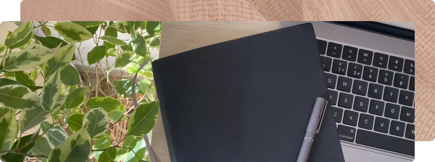 A laptop, black notebook, and pen on a wooden desk with a green leaf plant next to the desk.